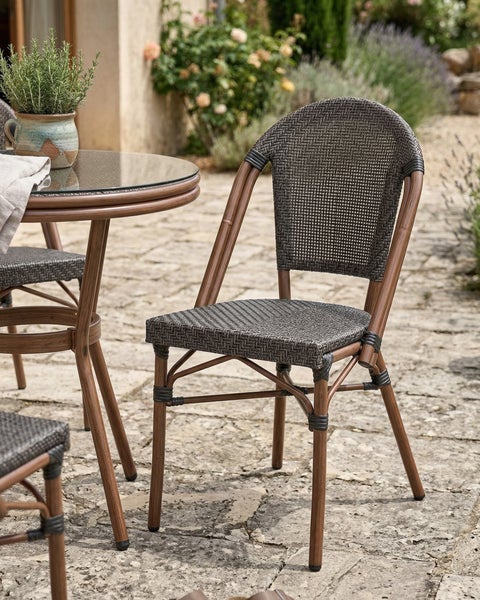 Bistro-Gartenstuhl mit geflochtener Sitzfläche und Gestell in Holzoptik neben passendem Tisch auf einer Steinterrasse.