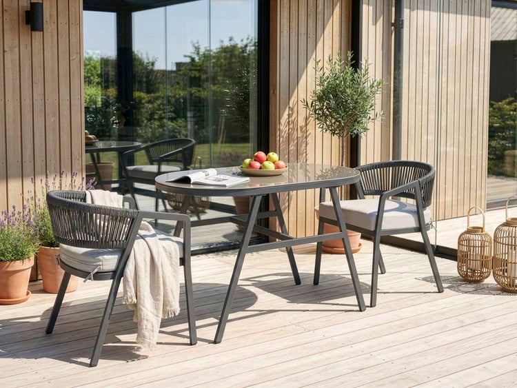 Gartenszene mit Esstisch, Stühlen, Pflanzen und Windlichtern auf einer Holzterrasse.