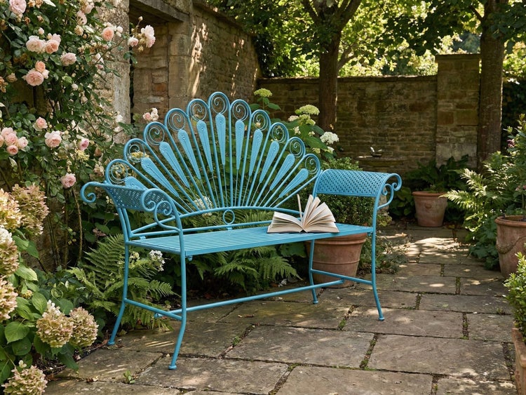 Blaue Gartenbank aus Metall mit Rückenlehne im Pfauendesign auf einer Steinterrasse inmitten von Rosen und grünen Pflanzen.