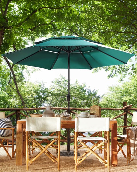 Großer Sonnenschirm über einem Esstisch aus Holz und Klappstühlen auf einer hellen Terrasse inmitten von Bäumen.