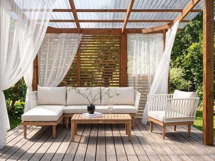 Moderne Gartenlounge unter Holzpergola mit lichtdurchlässigem Dach, hellen Polstern, Holztisch und weißen Vorhängen auf einer Holzterrasse.