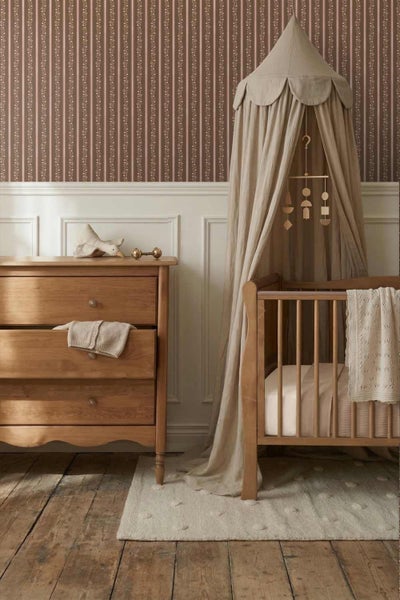 Kinderzimmer mit gestreifter Blumentapete, Holzkommode und Babybett mit Betthimmel.