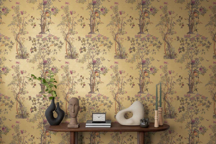 Vliestapete mit floralem Chinoiserie Muster und Baummotiven vor einer Konsole mit Dekorationsobjekten in einem Wohnraum.