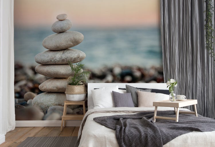 Vliestapete mit einem Motiv gestapelter Steine am Strand in einem hellen, modern eingerichteten Schlafzimmer mit Bett und Textilien.