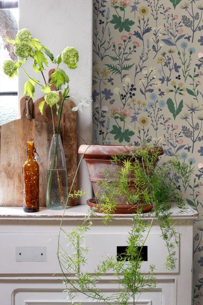 Helle Raumgestaltung mit floraler Tapete mit Wildblumenmuster hinter einer weißen Kommode mit Pflanzen und Holzbrettern.