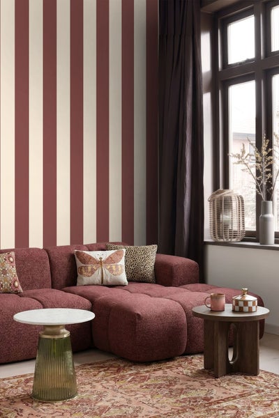 Gestreifte Tapete in einem Wohnzimmer mit rotem Sofa, gemustertem Teppich und kleinen Tischen. Vertikale Streifen in Rot und Beige.