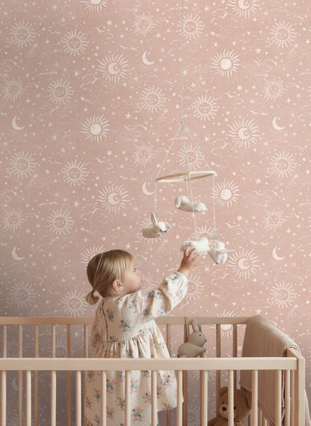 Kinderzimmer mit Tapete in Rosa mit weißen Sonnen, Monden, Sternen und Wolken, ein Kind im Holzgitterbett greift nach einem Mobile mit Stofftieren.