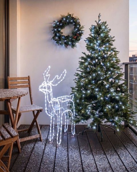 Weihnachtlich dekorierter Balkon mit beleuchtetem Weihnachtsbaum, LED-Rentier-Figur, Kranz und schneebedeckten Gartenmöbeln aus Holz.