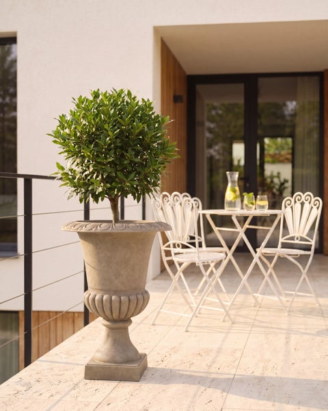 Verzierte Pflanzvase in Steinoptik mit einem rund geschnittenen Baum auf einer hellen Terrasse mit Bistromöbeln und Steinfliesen.