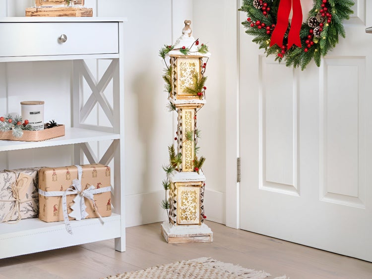 Dekorative Holzlaterne mit Winterdekoration neben einer weißen Tür mit Kranz und einem Regal mit Geschenken.