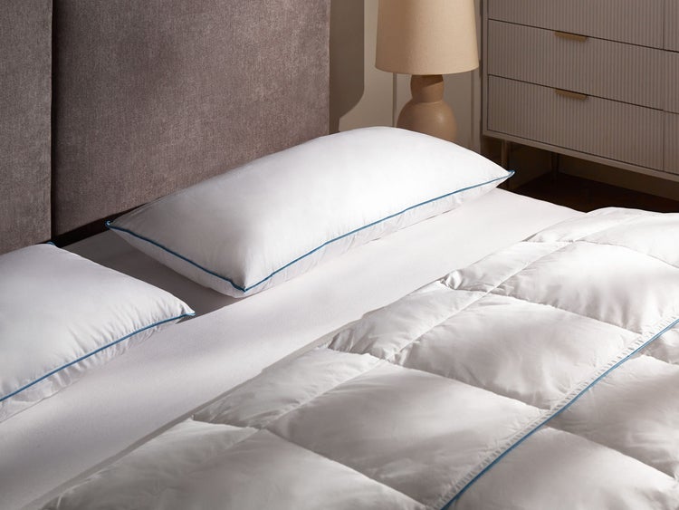 Weißes Kopfkissen und Steppdecke mit blauem Saum auf einem Bett in einem hellen Schlafzimmer mit grauem Kopfteil.