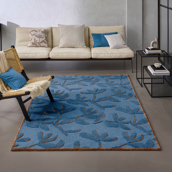 Blauer Teppich mit floralem Muster in einem modern gestalteten, hellen Wohnzimmer mit beigem Sofa, Korbsessel und schwarzem Beistelltisch.