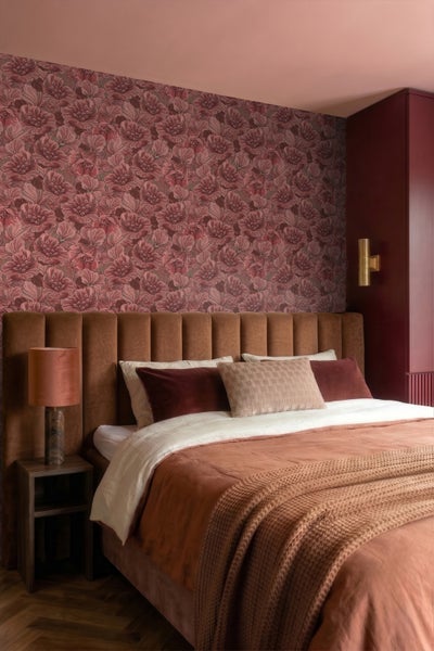Schlafzimmer mit floraler Mustertapete, gepolstertem Bett mit vertikal gestepptem Kopfteil, Nachttischlampe und warmen Textilien.
