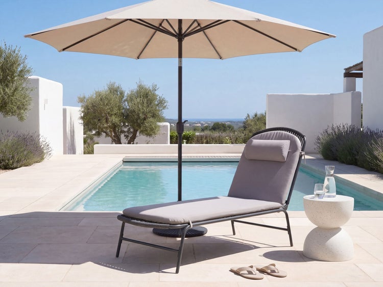 Gartenliege mit Polsterauflage und Kopfkissen unter einem Sonnenschirm auf einer Terrasse am Pool mit Beistelltisch und Flipflops.