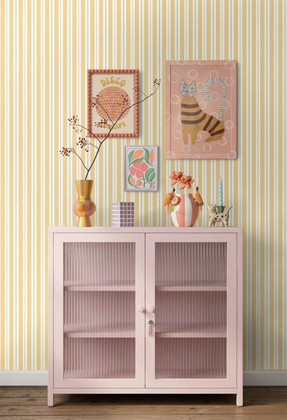 Vliestapete mit vertikalen Streifen in Ocker und Weiß hinter einer rosa Vitrine mit geriffeltem Glas und bunten Dekorationsartikeln.