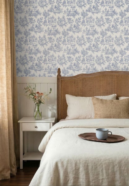 Schlafzimmer mit Toile de Jouy Tapete, Bett mit geflochtenem Kopfteil und Nachttisch mit Blumen in einer Vase.