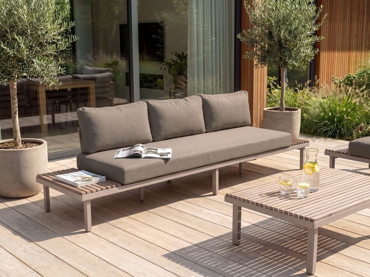 Modernes Loungesofa mit integrierten Ablageflächen auf einer Holzterrasse mit passendem Couchtisch und Olivenbäumen in Kübeln.