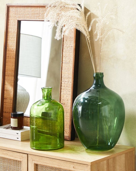 Dekorative Szene mit zwei grünen Glasvasen, getrocknetem Ziergras und einem Wandspiegel mit Rattanrahmen auf einer Holzkommode.