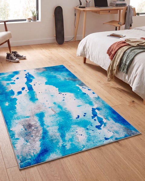 Moderner rechteckiger Teppich mit abstraktem Muster in Wasserfarben Optik liegend auf einem Holzboden in einem hellen Schlafzimmer.