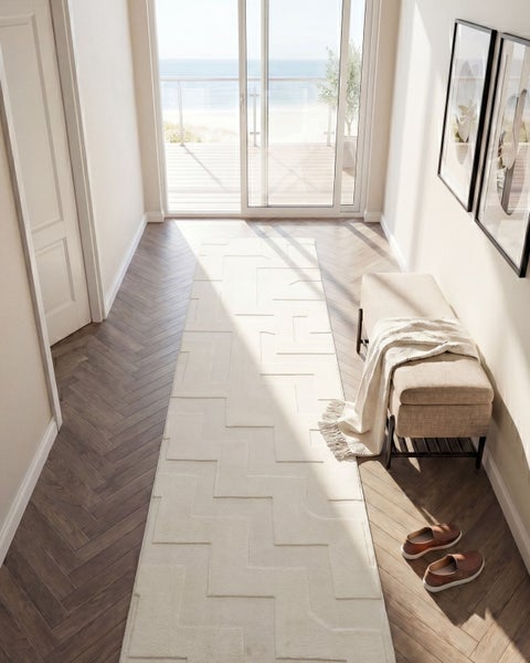 Ein heller Flur mit geometrischem Teppichläufer, einer Sitzbank mit Decke und Fischgrätparkett vor einer großen Terrassentür mit Meerblick.