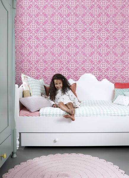 Ein Mädchen sitzt auf einem Bett vor einer Wand mit gemusterter Tapete in einem hellen Kinderzimmer.