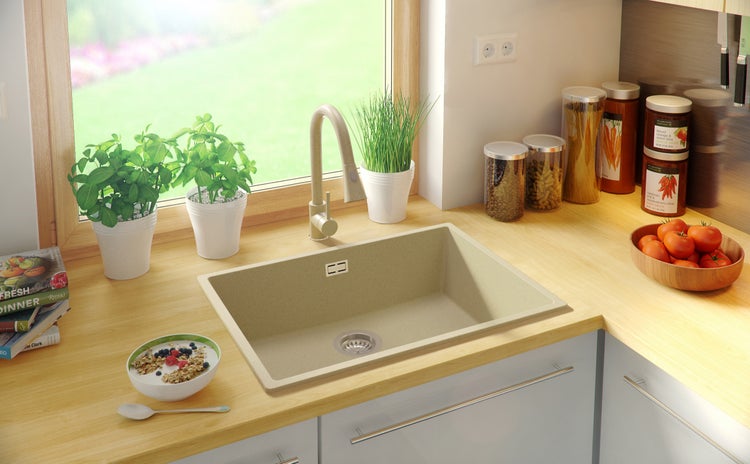 Küchenspüle in beiger Farbe mit Armatur, umgeben von Lebensmitteln und Pflanzen auf einer hellen Arbeitsplatte.