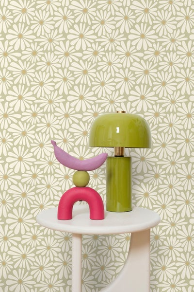 Vliestapete mit Gänseblümchen-Muster auf grünem Grund, moderne Szene mit Tischlampe und abstrakter Dekoration auf einem weißen Beistelltisch.