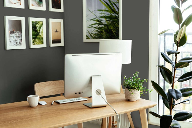 Moderner Arbeitsplatz mit hellen Holzmöbeln vor einer dunkelgrauen Wand mit weißen Bilderrahmen und Zimmerpflanzen.