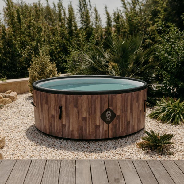 Aufblasbarer runder Whirlpool mit Außenverkleidung in Holzoptik auf einer Kiesfläche im hellen Garten, COCO SPA Logo.