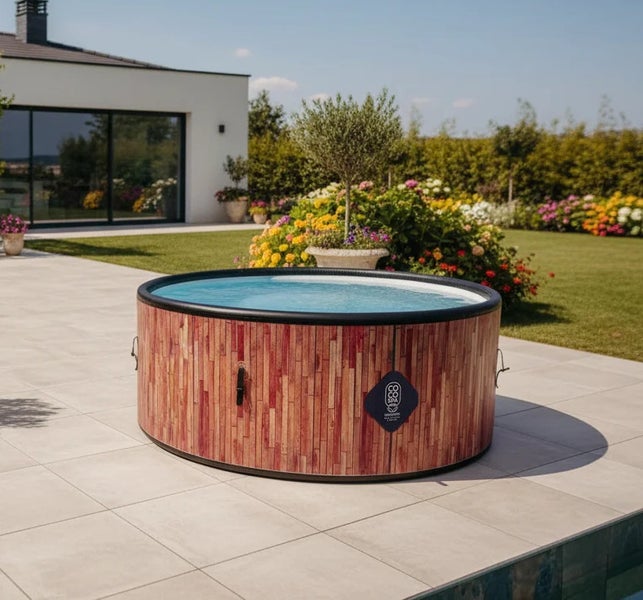 Aufblasbarer runder Whirlpool in Holzoptik auf einer hellen Fliesenterrasse in einem gepflegten Garten mit Blumenbeeten und CocoSpa Logo.