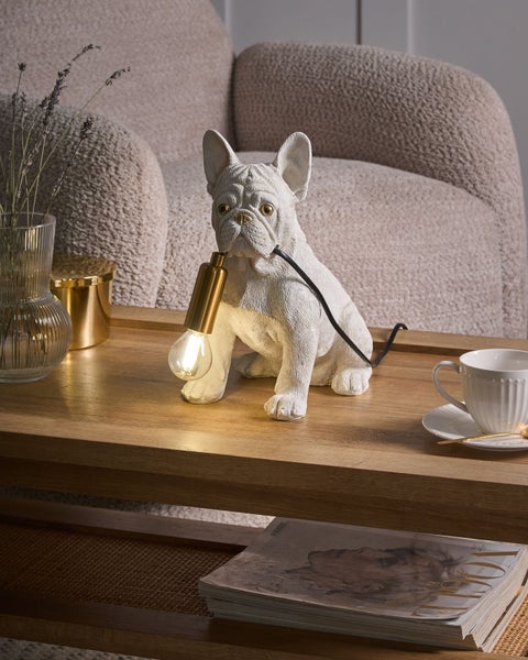 Dekorative Tischleuchte als weiße französische Bulldogge auf einem Holztisch in einem hellen Wohnzimmer neben einem beigefarbenen Sofa.