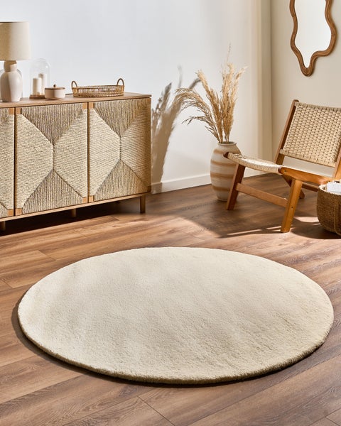 Runder, weicher cremefarbener Teppich auf Holzboden in hellem Wohnzimmer mit Holzkommode, Relaxsessel und Naturdekoration.