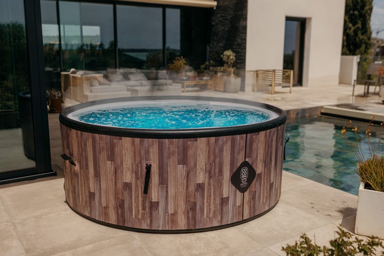 Runder aufblasbarer Whirlpool in Holzoptik mit blauem Wasser und Coco Spa Logo, aufgestellt auf einer Terrasse neben einem Swimmingpool.