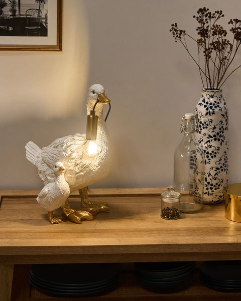 Tischleuchte in Form einer Gans, die ein Leuchtmittel im Schnabel hält, mit kleiner Gösselfigur auf einem Holzsideboard.