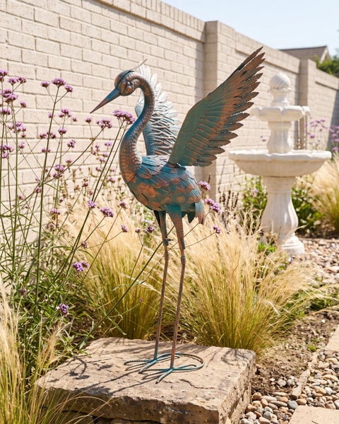 Metall-Gartenfigur Reiher mit ausgebreiteten Flügeln in einem hellen Garten mit Gräsern und Springbrunnen.