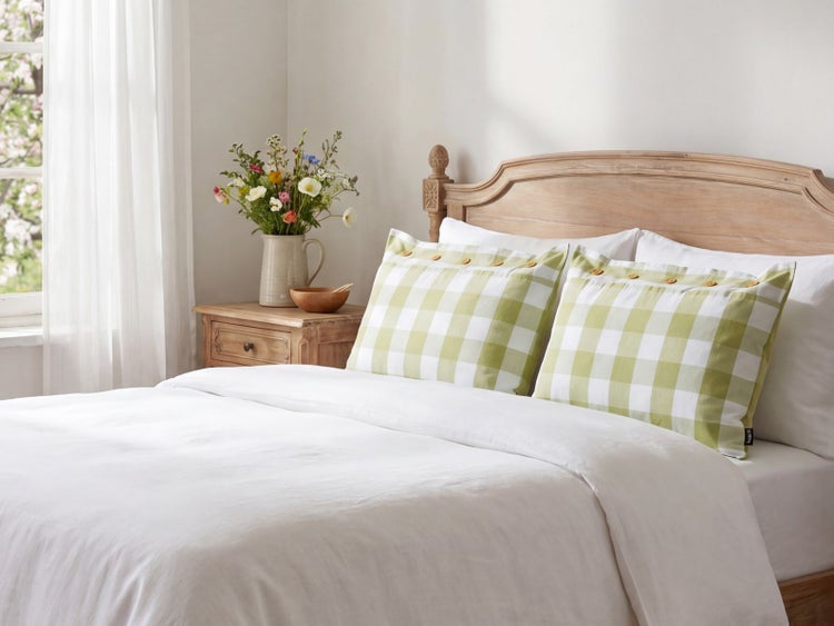 Schlafzimmer im Landhausstil mit Holzbett, Bettwäsche, zwei karierten Kissen mit Knöpfen und Blumenstrauß auf dem Nachttisch.
