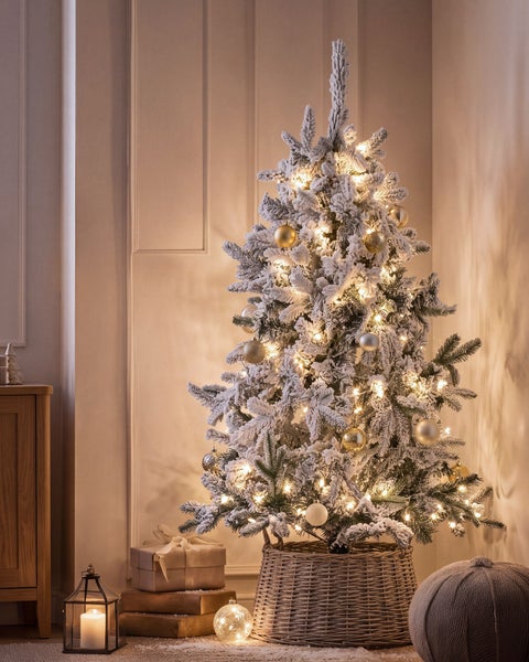 Ein künstlicher, verschneiter Weihnachtsbaum steht in einem geflochtenen Korb in einem weihnachtlich geschmückten Raum mit Lichterkette.