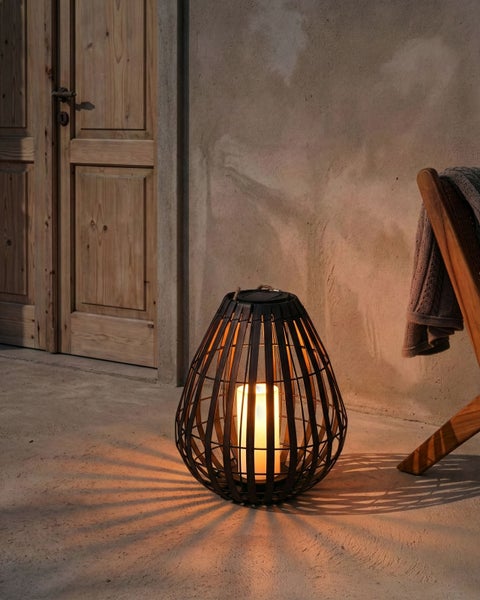 Solar-Laterne aus dunklem Bambusgeflecht mit LED-Kerze auf einer Terrasse neben einer Holztür, wirft dekorative Schatten.