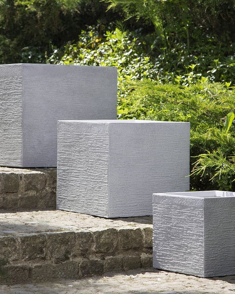 Drei graue, würfelförmige Pflanzkübel aus Stein im Garten.