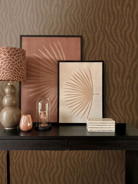 Stilvolles Interieur mit gemusterter Tapete, einem Tisch, zwei gerahmten Bildern mit Palmblattmotiven, einer Tischlampe und Dekorationsartikeln.