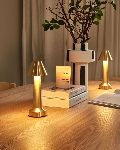 Dekoratives Arrangement mit zwei goldenen Tischleuchten, einer Kerze, Büchern und einer Vase mit Zweigen auf einem Tisch.