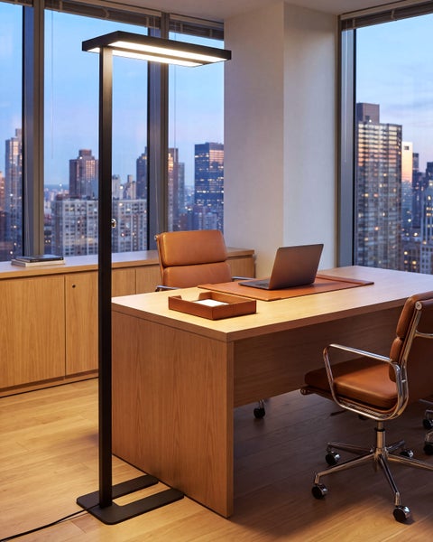 Moderne Stehlampe mit flachem Leuchtkopf in einem elegant eingerichteten Büro mit Schreibtisch und Blick auf die Skyline bei Dämmerung.