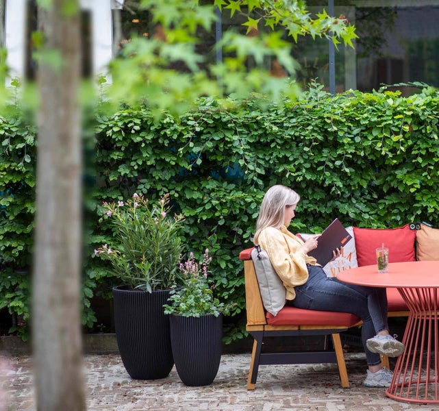 Frau auf einer Gartenbank mit Polstern neben zwei hohen geriffelten Pflanzkübeln und einem runden Tisch vor einer Hecke.