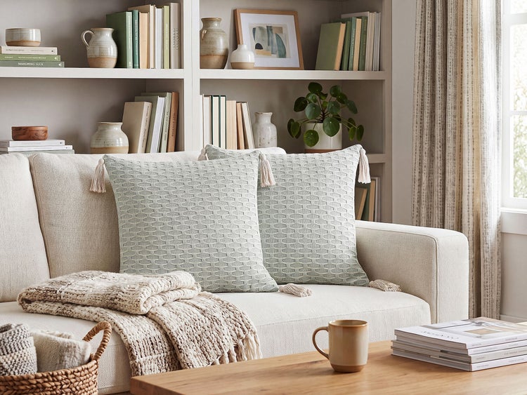 Zwei quadratische Sofakissen mit Strukturmuster und Quasten auf einem hellen Sofa vor einem Bücherregal in einem wohnlich eingerichteten Raum.