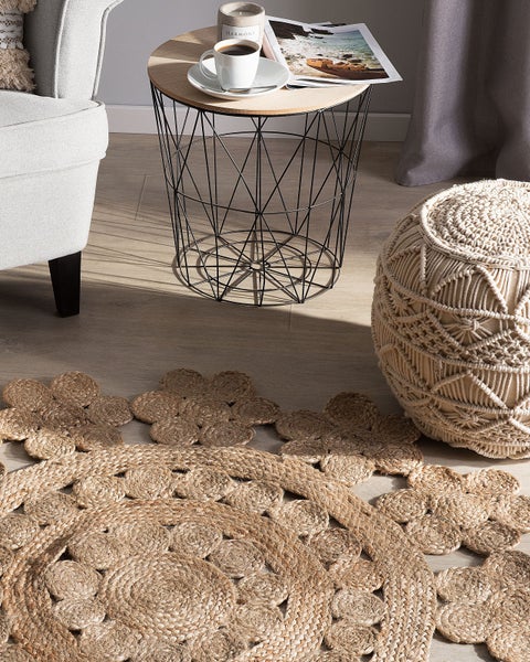 Helle Wohnzimmerszene mit rundem Juteteppich in Blumenform, Beistelltisch aus Metalldraht, Makramee-Pouf und Sessel auf Holzboden.
