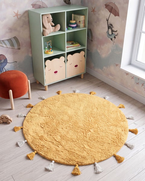 Runder Kinderteppich mit Troddeln in einem Kinderzimmer mit Regal und Sitzhocker.