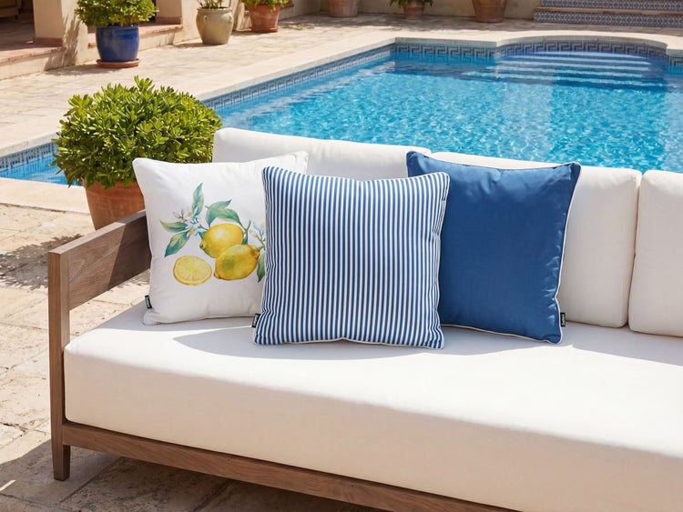 Drei dekorative Gartenkissen auf einem Holzsofa am Pool, eines mit Zitronenmuster, eines gestreift und eines unifarben.