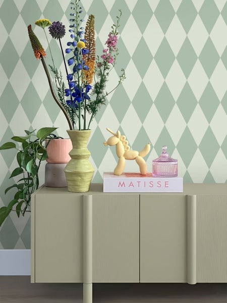 Dekoratives Wohnzimmerambiente mit Blumenvase, Ballon-Einhorn und Buch auf einer Kommode vor einer Rautenmuster-Tapete.