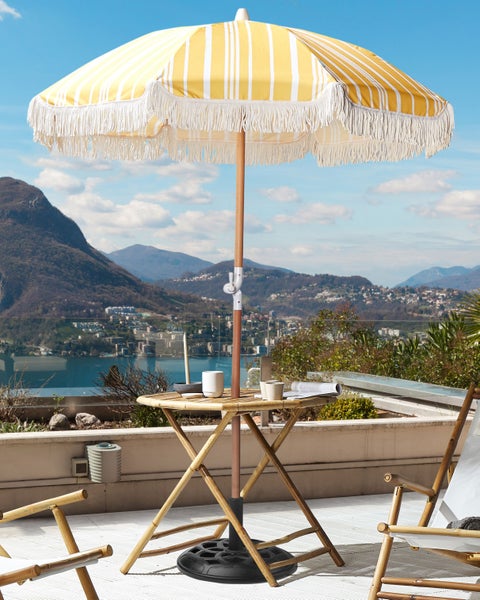 Sonnenschirm mit gelb-weißen Streifen und Fransen über einem klappbaren Bambustisch auf einer Terrasse mit See- und Bergblick.