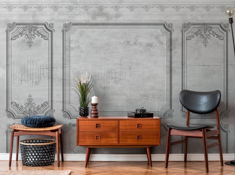 Fototapete mit klassischer Stuck-Optik in Grau, kombiniert mit einem Holz-Sideboard, Stuhl und Hocker in einem hellen, modern eingerichteten Raum.